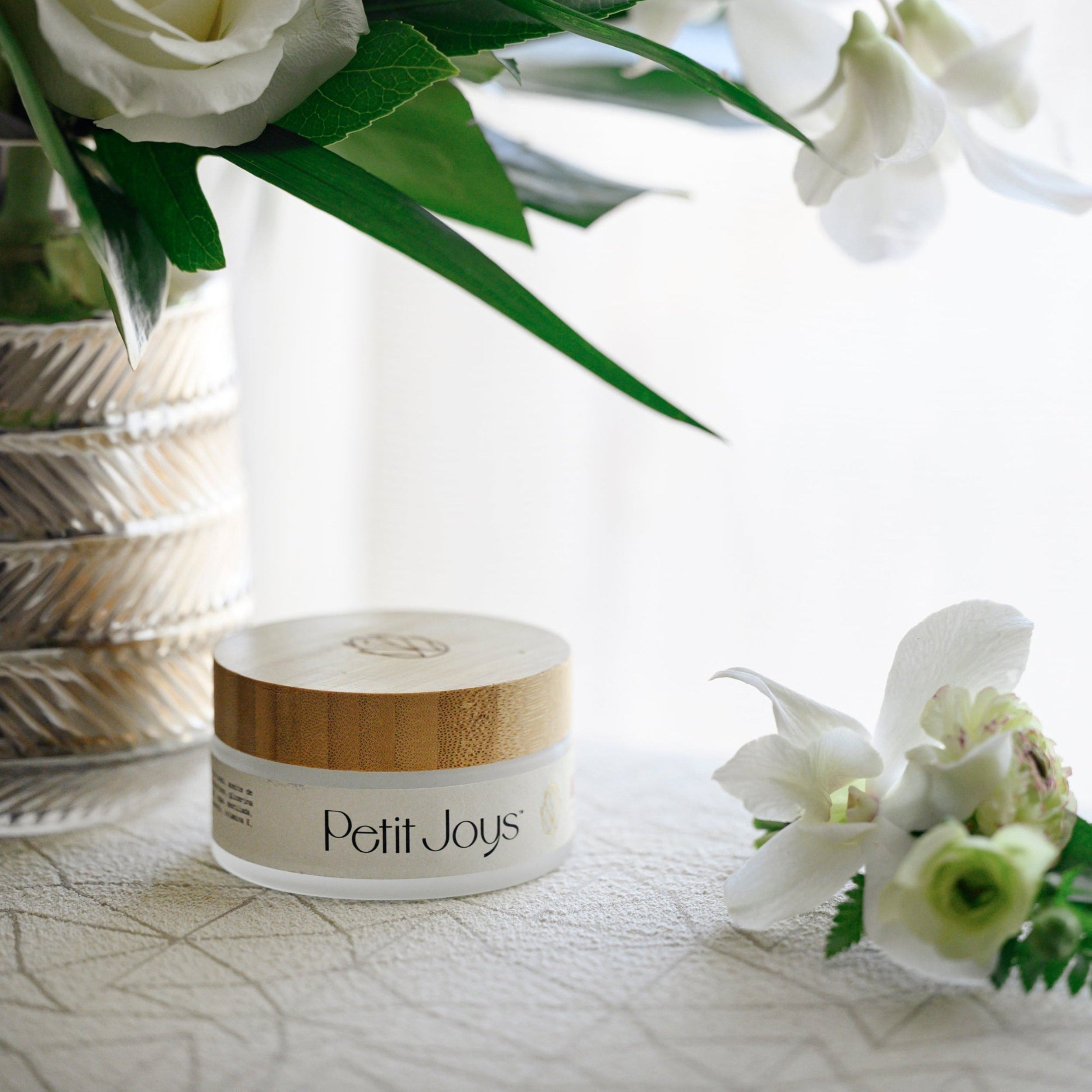 Body Butter jar of Petit Joys cream with white flowers and green leaves on a light background
